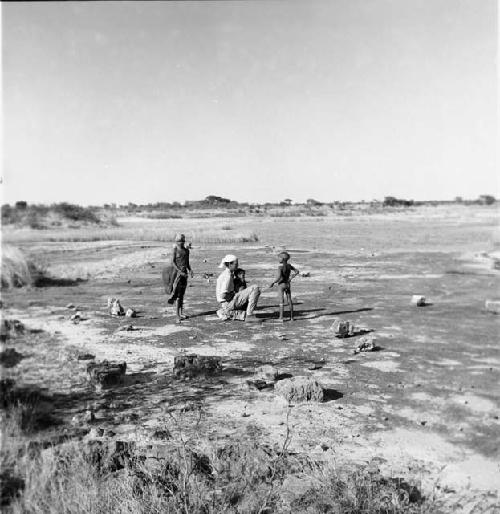 Children standing next to Robert Gardner sitting on a rock near edge of a pan, holding child on his lap