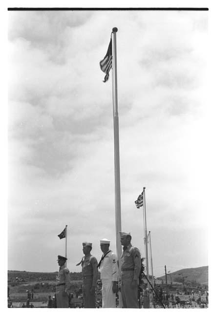 Military personnel of various groups standing in front of raised flags