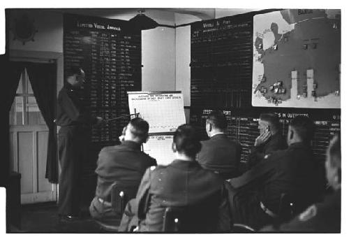 American military personnel reviewing port and cargo charts; central chart reads: "AVERAGE TIME DISCHARGING AND BACKLOADING OF DEEP DRAFT CARGO VESSELS"