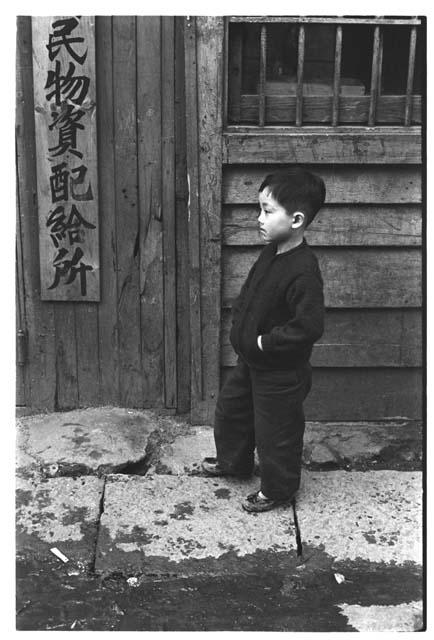 Young Korean boy outside building with vertical sign on door; sign indicates government agency providing indispensable supplies during an emergency situation