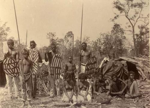 Large group of Aboriginal people, men and boys standing, holding spears and shields, and women seated on the ground