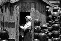 Woman in doorway of wooden shed; piles of jars to left and right