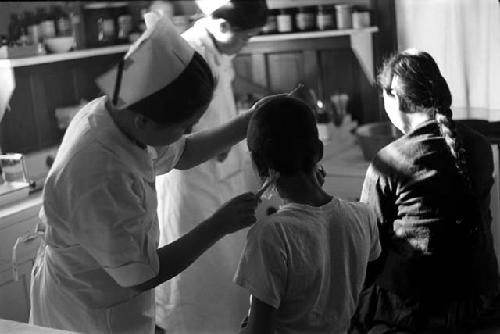 Nurse examining ear of patient; another nurse and patient in background.