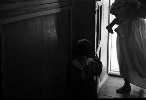 Door ajar, letting in sunlight; woman in doorway holding infant.