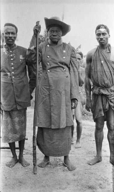 Chief with staff and other men wearing military jacket over traditional clothing