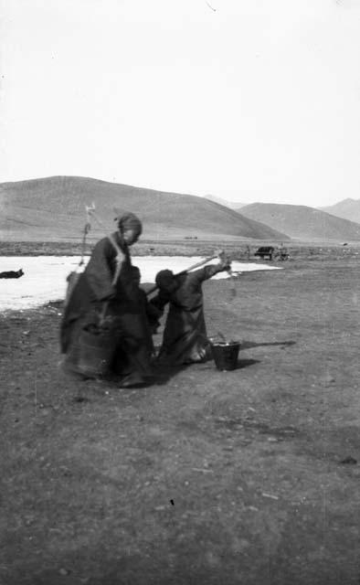 Two women fetching water using buckets on shoulder, pole