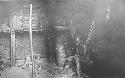 Individual sitting by hearth of hut with pots, baskets, house goods