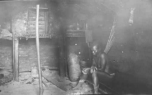 Individual sitting by hearth of hut with pots, baskets, house goods