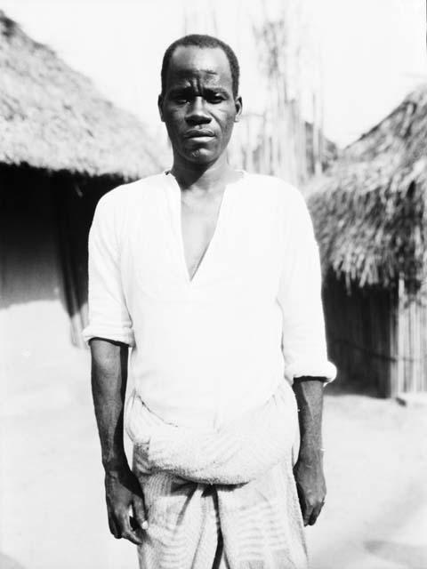 Man standing in pants and shirt in front of hut, front view