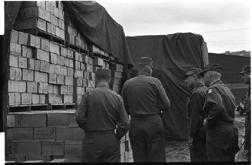 Military personnel standing beside a pile of boxes