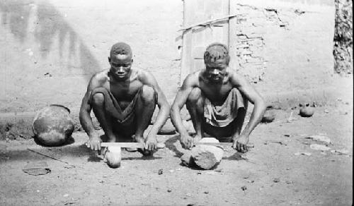 Two men sitting on ground sharpening cutlasses or machetes on rock