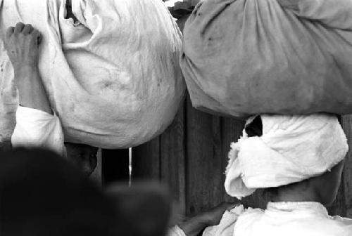 Two women holding large cloth wrapped bundles on their heads.