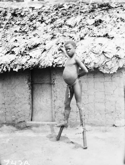 Young boy on stilts in front of hut