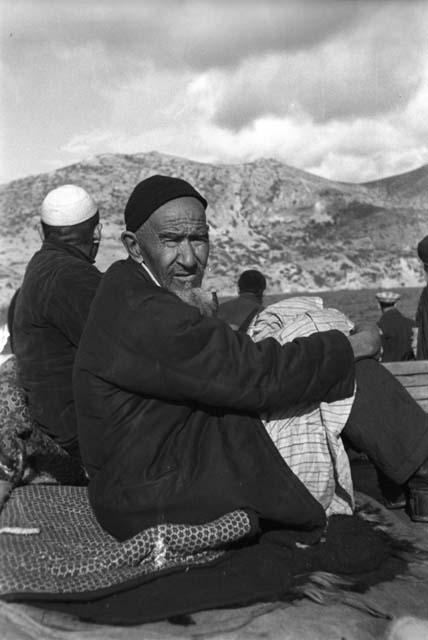 Bearded man, on boat, more people behind him, mountains in background