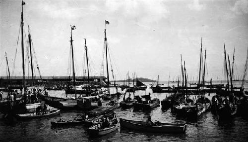 Men in rowboats and boats with masts in port