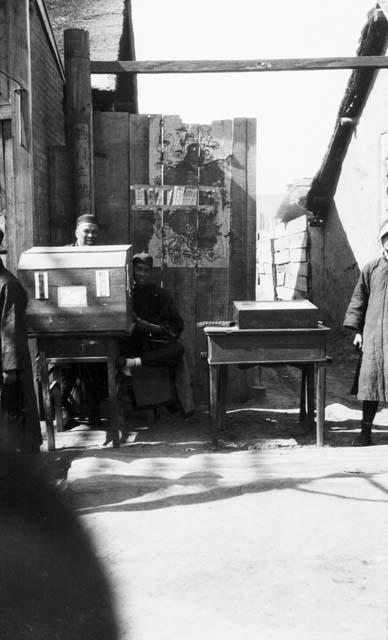 Public letter writer, two men behind a desk with poster on wall behind