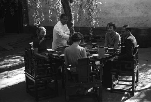 Eleanor Lattimore, two women and one man all seated at round table in courtyard