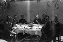 Eight men seated at round table in courtyard