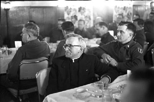 Priest and men sitting at tables looking ahead