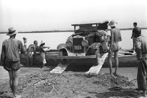 Group of men, some naked, unloading car from raft along two planks