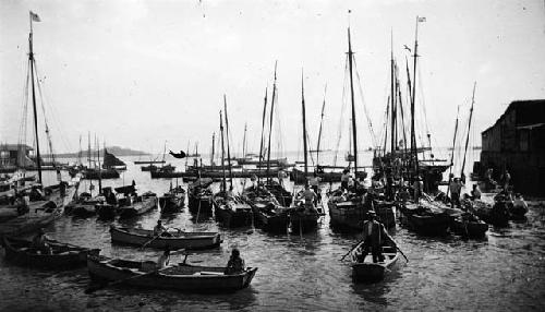 Men and woman in rowboats and single-mast boats in port