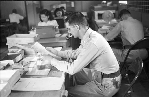 Soldier sitting at desk smoking pipe while reading, with typing pool in the background