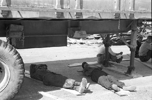 Men laying on wooden planks under truck avoiding the sun