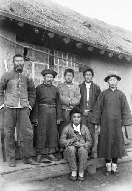 Five men standing, one sitting holding a cat, on step in front of building