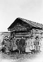 Several women and children, seated, in front of a wooden building