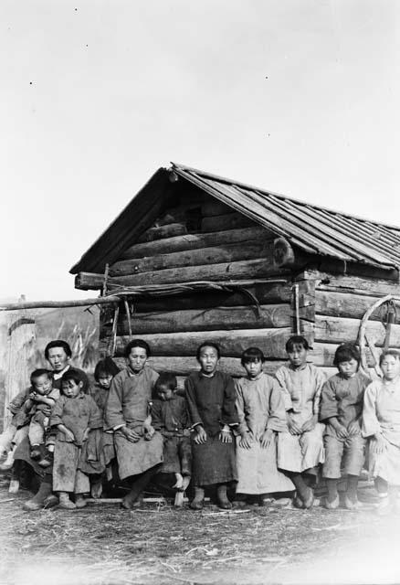 Several women and children, seated, in front of a wooden building