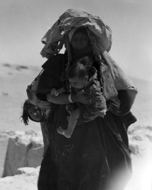 Shammar Bedouin at Jumaima, woman carrying load on head and baby in her arms