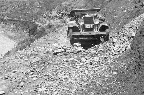 Car on rocky hillside road, alongside body of water