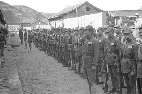 Two long rows of soldiers, buildings and wall in background