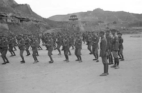 Soldiers at drill practice. Cliff, city gate, buildings in distance