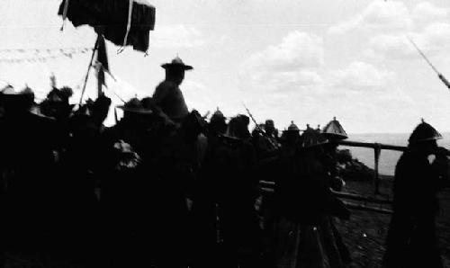 Crowd of people in pointed hats carrying ceremonial umbrella and man on chair