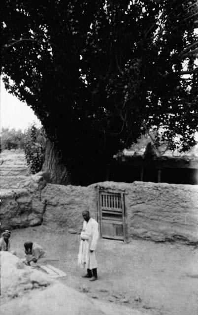 Two men and woman in area inside mud-brick wall with wooden gate