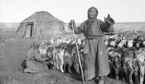 Woman kneeling milking a goat, and older woman holding a staff