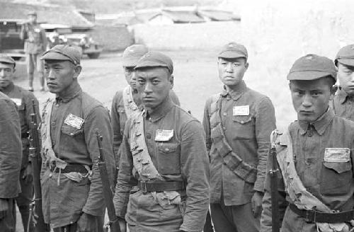 Close-up of two rows of soldiers. Buildings, a wall and car in background