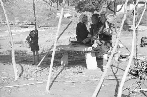 Three women sitting on a bench in a shaded area. Child nearby, buildings behind