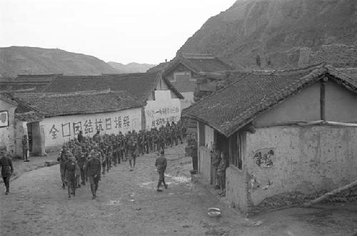 View of long line of soldiers marching along road through the village