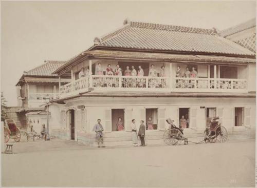 Women standing on balcony, in windows and men standing in front of building