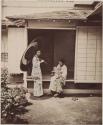 One woman standing, one woman sitting and breastfeeding child