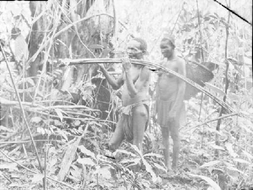 Woman with basket following man carrying reeds through jungle