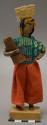 Doll, woman with basket on head and roll of plant fiber in arm, affixed to stand