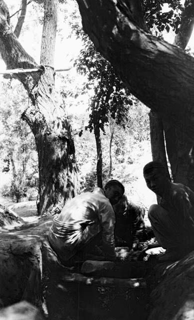 Chantos at fire, three men crouched around something under a large tree