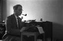 Nat Peffer at typewriter, smoking a pipe with desk behind him