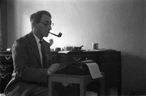 Nat Peffer at typewriter, smoking a pipe with desk behind him