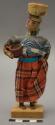 Doll, woman with basket on head and cup in arm, affixed to stand