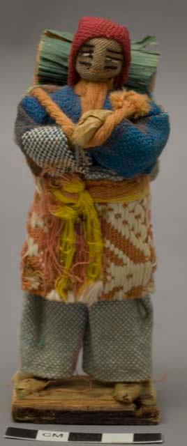 Doll, man with cloth bag and bundle on back, affixed to stand