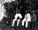 Three men sitting on bench with horses in background